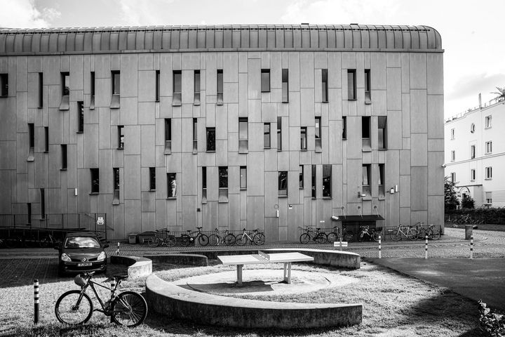 Schwarz-Weiß-Foto eines Innenhofs, in deren Vordergrund eine Tischtennisplatte von runden Sitzbänken umgeben ist, im Hintergrund ragt ein moderner Bau in die Höhe mit schmalen, langen Fenstern und vielen Fahrrädern an der Hauswand
