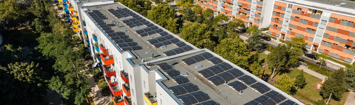 Luftaufnahme eines Mehrfamilienhauses mit Solaranlagen auf dem Dach, bunten Balkonen und grüner Umgebung.