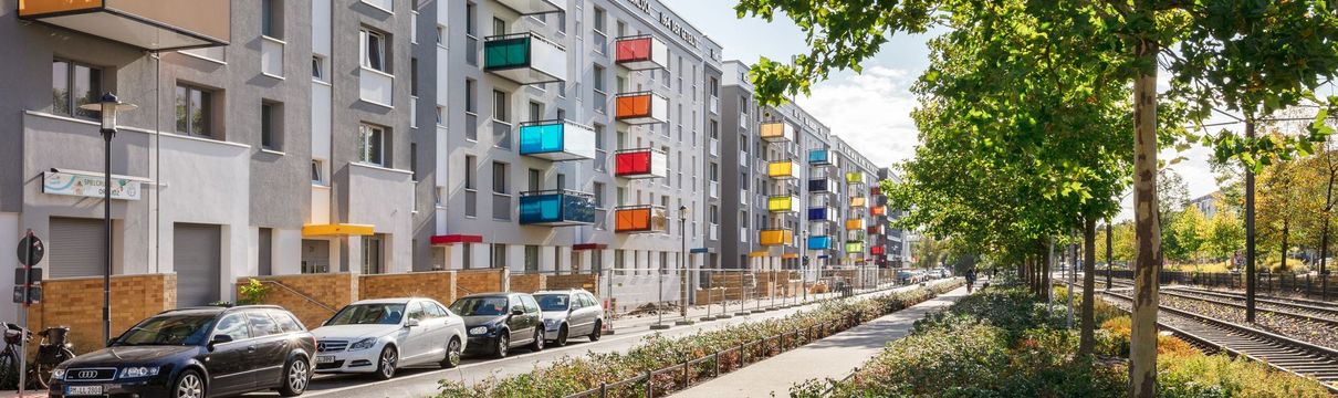 Moderne Wohngebäude mit farbigen Balkonen entlang einer begrünten Straße mit Bäumen und Straßenbahngleisen.