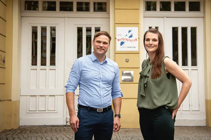 Ein Mann mit hoch gekrempelten Ärmeln in hellblauem Hemd und eine brünette Frau in dunkelgrüner Bluse stehen lächelnd vor den weißen Eingangstüren eines gelben Gebäudes