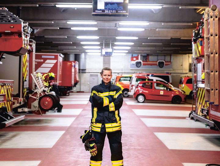 Junge Feuerwehrfrau steht im Innern einer Feuerwehrwache zwischen den Einsatzfahrzeugen