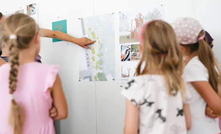 Kinder betrachten eine Wand mit Plänen und Bildern, während eine Person auf einen Lageplan zeigt.