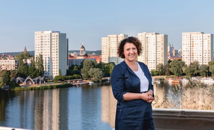 Potsdams Sozialbeigeordnete Brigitte Meier steht lächeln in dunkelblauem Kostüm vor einer Potsdamer Skyline, die die Havelbucht zeigt.