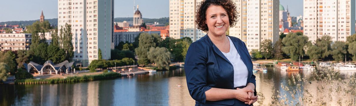 Potsdams Sozialbeigeordnete Brigitte Meier steht lächeln in dunkelblauem Kostüm vor einer Potsdamer Skyline, die die Havelbucht zeigt.