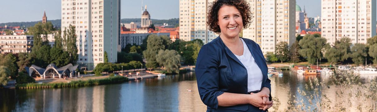 Potsdams Sozialbeigeordnete Brigitte Meier steht lächeln in dunkelblauem Kostüm vor einer Potsdamer Skyline, die die Havelbucht zeigt.