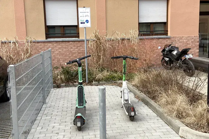 Zwei E-Scooter stehen an einer markierten Abstellfläche vor einem Wohngebäude, daneben ein Motorrad.