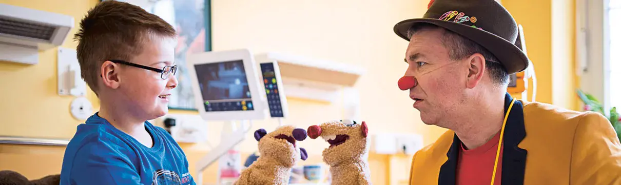Kind in blauem Pullover mit Brille auf einem Krankenhausbett sitzend spielt mit seiner Handpuppe zusammen mit der Handpuppe eines Clowns, der am Krankenbett sitzt
