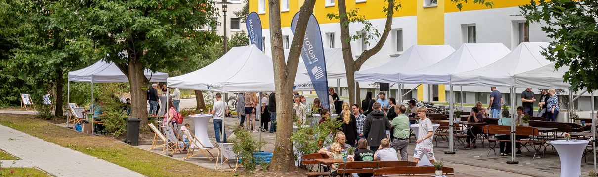 Festliche, weiße Pavillons und hölzerne Sitzgelegenheiten mit vielen Menschen vor einem Plattenbau mit gelben Streifen