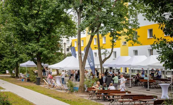 Festliche, weiße Pavillons und hölzerne Sitzgelegenheiten mit vielen Menschen vor einem Plattenbau mit gelben Streifen