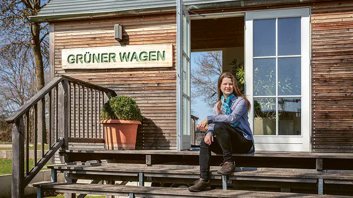 Eine Frau sitzt auf einer Holztreppe in der Sonne, die Treppe gehört zu einem hölzernen Bauwagen, am Bauwagen ist ein Schild mit Grüner Wagen zu sehen
