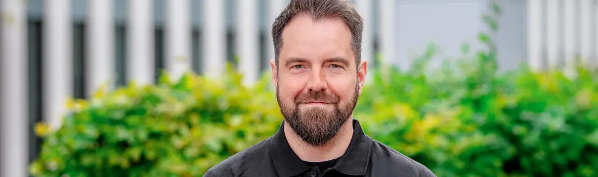 Portrait einer Person im schwarzen ProPotsdam-Poloshirt vor grüner Hecke und modernem Gebäude im Hintergrund