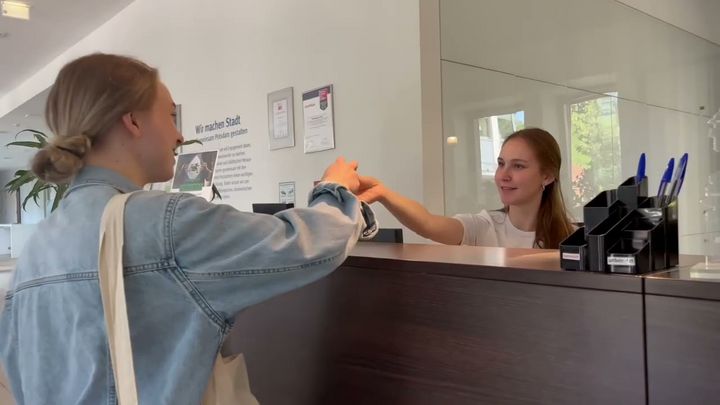 Arbeitsplatz am Fenster mit einem fest installiertem, laufendem Computer und einem offenen Laptop, an dem eine junge Frau mit langen, blonden Haaren sitzt, die etwas oder jemanden neben ihrem Arbeitsplatz anzuschauen scheint.