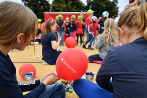 Zwei Mädchen im Schneidersitz sitzen auf dem Boden. Das eine Mädchen hält einen roten Luftballon mit der Aufschrift Sanierungsträger Potsdam in der Hand. Im Hintergrund weitere Kinder.