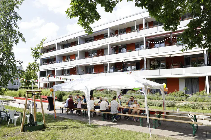 Wohnhaus mit durchgezogenen Balkonflächen vor dem Menschen im Grünen unter Sonnensegel beisammen sitzen