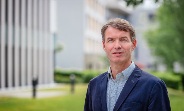 Porträt eines Mannes mit grau melierten, kurzen Haaren, hellem, gemusterten Hemd und blauem Sommerjacket. Im Hintergrund sind verschwommen Gebäudestrukturen wahrnehmbar.