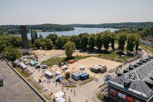 Luftaufnahme eines Stadtteilfests in Potsdam-Krampnitz an einem sonnigen Tag. Auf einer großen, offenen Fläche sind mehrere weiße Zelte und Stände aufgebaut, zwischen denen sich viele Besucherinnen und Besucher bewegen. Ein gepflasterter Rundweg führt um das Gelände, auf dem sich auch Menschen mit Fahrrädern aufhalten. Rechts im Bild steht ein mehrstöckiges Gebäude im Bau mit Gerüst und Bannern, links ein hoher, historischer Backsteinturm inmitten von Bäumen. Im Hintergrund erstreckt sich ein weiter See mit kleinen Booten, umgeben von dichtem Wald.