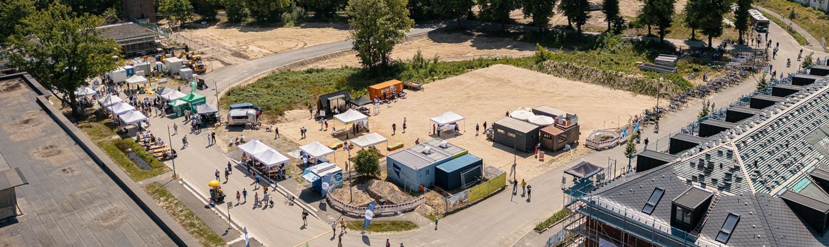 Luftaufnahme eines Stadtteilfests in Potsdam-Krampnitz an einem sonnigen Tag. Auf einer großen, offenen Fläche sind mehrere weiße Zelte und Stände aufgebaut, zwischen denen sich viele Besucherinnen und Besucher bewegen. Ein gepflasterter Rundweg führt um das Gelände, auf dem sich auch Menschen mit Fahrrädern aufhalten. Rechts im Bild steht ein mehrstöckiges Gebäude im Bau mit Gerüst und Bannern, links ein hoher, historischer Backsteinturm inmitten von Bäumen. Im Hintergrund erstreckt sich ein weiter See mit kleinen Booten, umgeben von dichtem Wald.