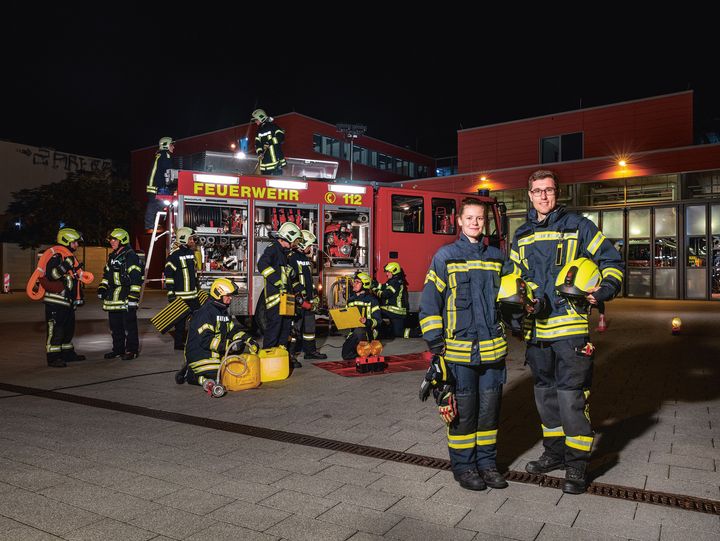 Eine Feuerwehrfrau und ein Feuerwehrmann stehen vor einem Feuerwehrfahrzeug bei dem weitere Feuerwehrleute tätig sind