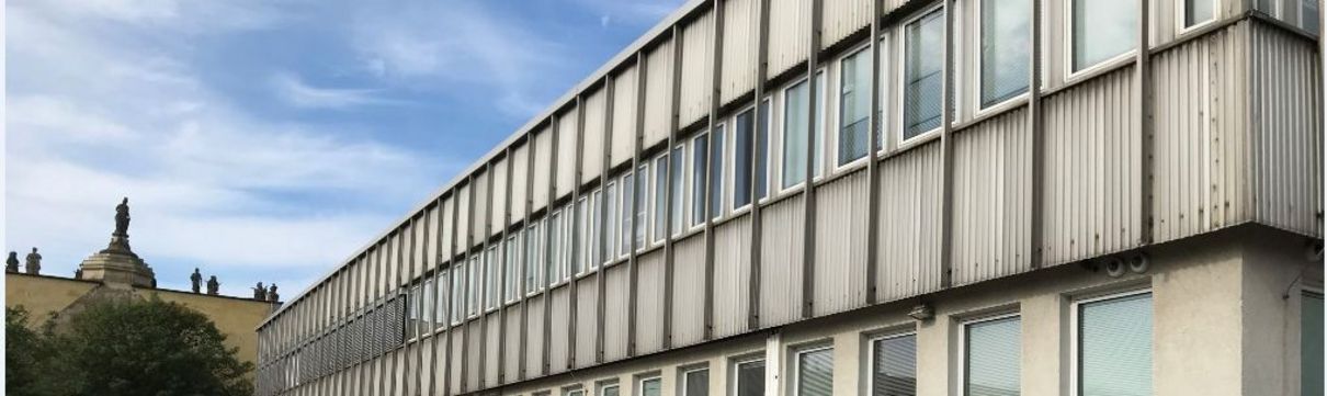 Ein zweistöckiger Flachbau mit hellgrauer Fassade, vor dem ein paar Autos parken.