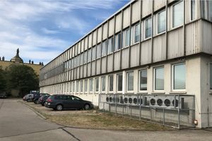 Ein zweistöckiger Flachbau mit hellgrauer Fassade, vor dem ein paar Autos parken. 