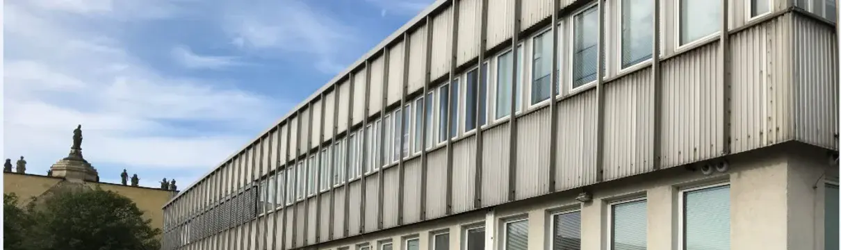 Ein zweistöckiger Flachbau mit hellgrauer Fassade, vor dem ein paar Autos parken.