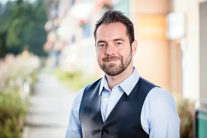Mike Didschun blickt in die Kamera. Im Hintergrund sind schemenhaft ein Haus und ein Fußweg zu sehen.