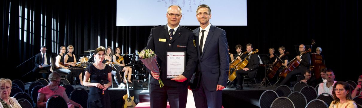 Zwei Männer, von denen einer Blumen und eine Urkunde hochhält, in dunklen Anzügen stehen vor einer Orchesterbühne im Zuschauergang und blicken in die Kamera