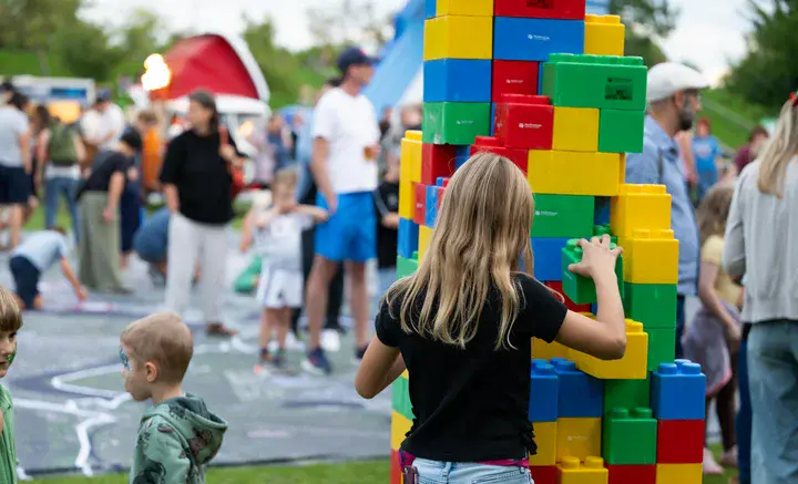 Kind baut bei einem Familienfest mit großen bunten Bausteinen einen Turm, Besucherinnen und Besucher im Hintergrund.