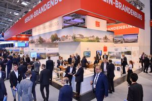 Männer und Frauen in Anzügen an einem Messestand und auf den Gängen einer Messehalle