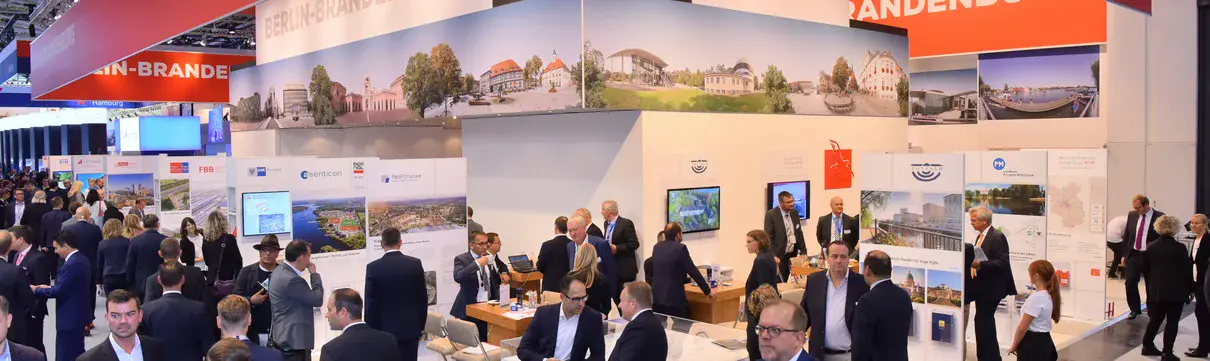 Männer und Frauen in Anzügen an einem Messestand und auf den Gängen einer Messehalle