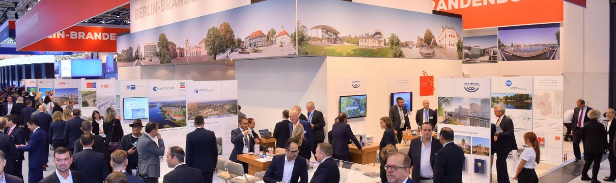 Männer und Frauen in Anzügen an einem Messestand und auf den Gängen einer Messehalle