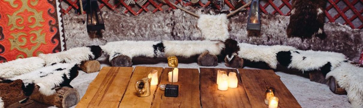 In einer Nomadenjurte steht ein Tisch mit Kerzen und kleinen Figuren, dazu mit Fellen überzogene Sitzgelegenheiten.