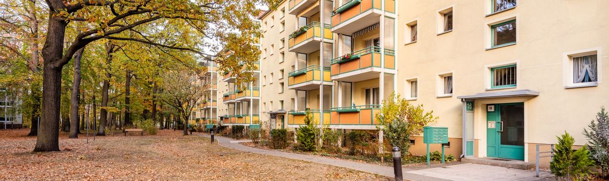 herbstliche Bäume, an denen sich ein Weg entlang eines Plattenbaus mit gelben und orangen Balkonen entlang schlängelt