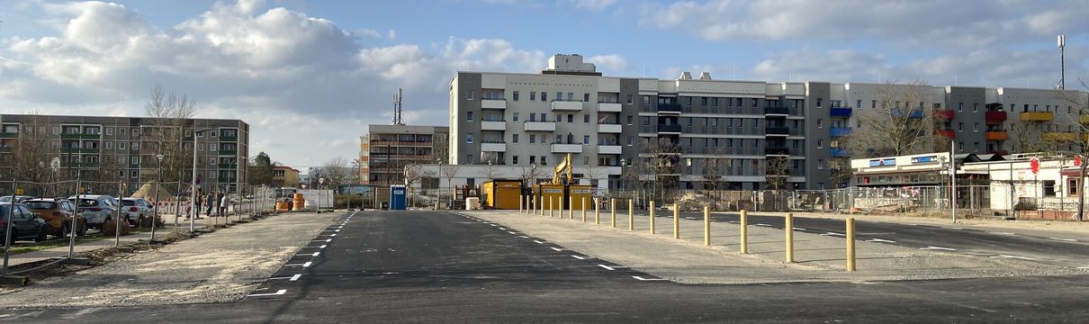 Asphaltierte Parkplatzfläche mit Gebäuden im Hintergrund und blauem Himmel.