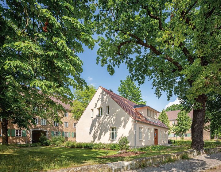 Seitenansicht eines zweietagigen Häuschens im Grünen, welches im Hintergrund von einem größeren, hellem Backsteinbau umrahmt wird