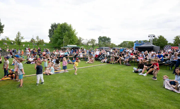 Großes Familienfest im Park mit zahlreichen Besucherinnen und Besuchern auf einer Wiese vor Sitzbänken und Bühne.