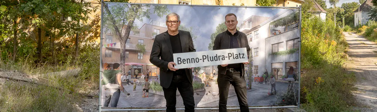 Zwei Männer stehen mit einem Straßenbild vor einem Haus und einer Tafel mit einem Bild.