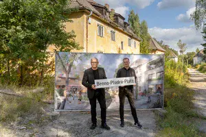 Zwei Männer stehen mit einem Straßenbild vor einem Haus und einer Tafel mit einem Bild.