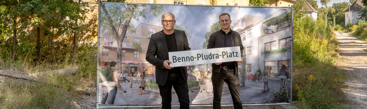 Zwei Männer stehen mit einem Straßenbild vor einem Haus und einer Tafel mit einem Bild.