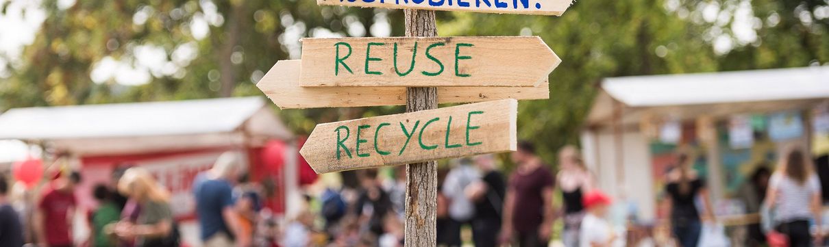 Ein aus Holz gefertigter Wegweiser, auf dem die Wärter Ausprobieren, Reuse und Recycle stehen. Im Hintergrund sind Menschen zu sehen, die an Informations- und Ausstellungsständen stehen.