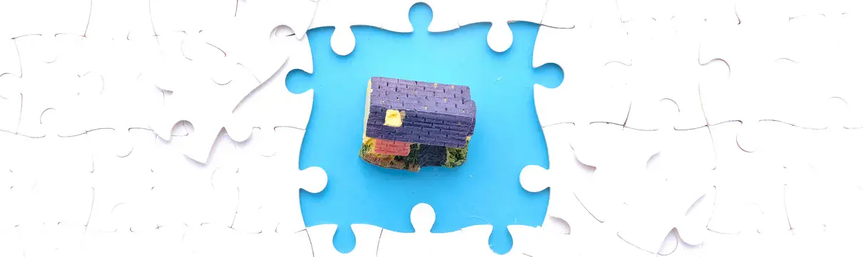 Draufsicht auf weißes Puzzle bei denen in der Mitte vier Puzzleteile fehlen, die ein Grundstück mit Haus freigegeben