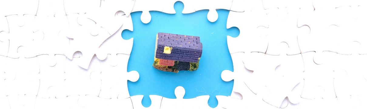 Draufsicht auf weißes Puzzle bei denen in der Mitte vier Puzzleteile fehlen, die ein Grundstück mit Haus freigegeben