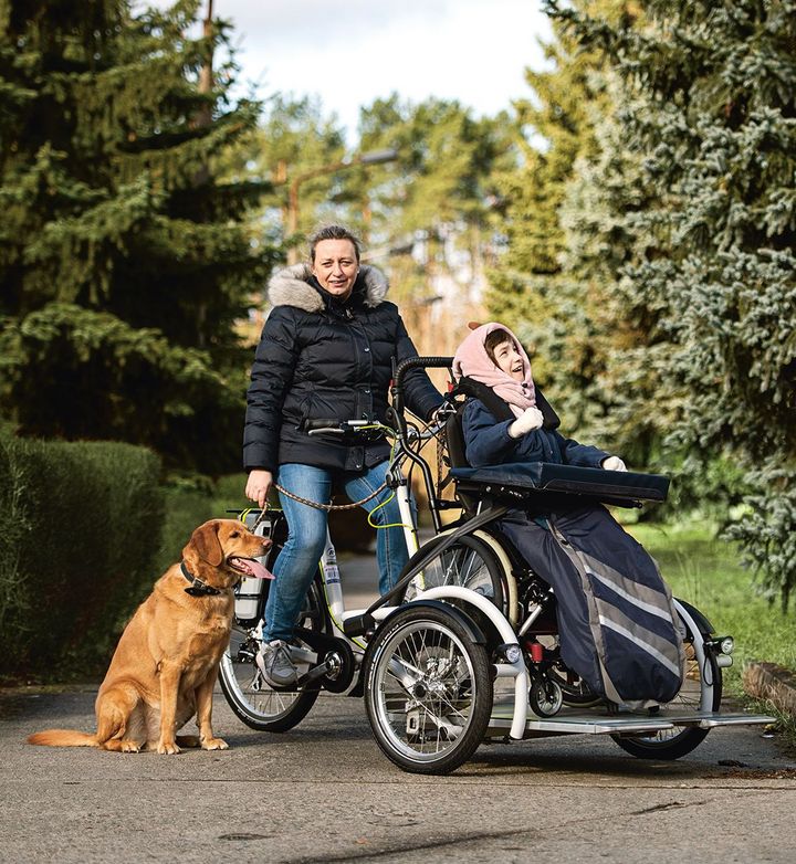 Zu sehen sind eine Mutter mit ihrer im Rollstuhl sitzenden Tochter auf einem Lastenra. Mit dabei ist ein Hund, im Hintergrund sind Bäume zu sehen.