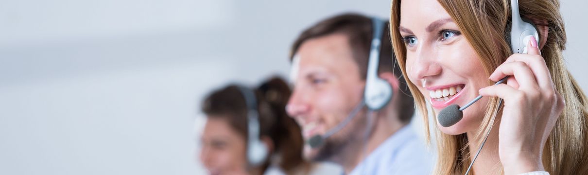 Drei Köpfe im Profil sind von Personen zu sehen, die nebeneinander sitzen, ein Headset tragen und telefonieren. Die vordere Person, eine junge Frau steht im Fokus, die anderen zwei Personen sind unscharf weiter hinten im Bild.
