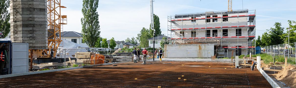 Eine Gruppe von Menschen steht auf einer freigeräumten Baufläche auf einer Baustelle. Es sind Baukräne und ein mit einem Baugerüst verkleidetes Gebäude im Hintergrund zu sehen.