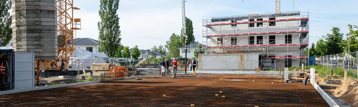 Baustart für die neue Kita in der Gartenstadt Nord im Bornstedter Feld (Fotograf: Benjamin Maltry) Eine Gruppe von Menschen steht auf einer freigeräumten Baufläche auf einer Baustelle. Es sind Baukräne und ein mit einem Baugerüst verkleidetes Gebäude im Hintergrund zu sehen.
