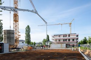 Eine Gruppe von Menschen steht auf einer freigeräumten Baufläche auf einer Baustelle. Es sind Baukräne und ein mit einem Baugerüst verkleidetes Gebäude im Hintergrund zu sehen.