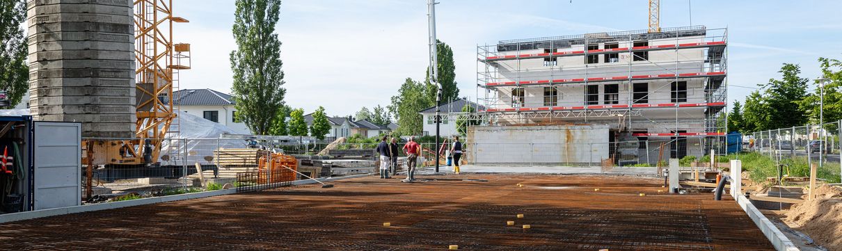 Eine Gruppe von Menschen steht auf einer freigeräumten Baufläche auf einer Baustelle. Es sind Baukräne und ein mit einem Baugerüst verkleidetes Gebäude im Hintergrund zu sehen.