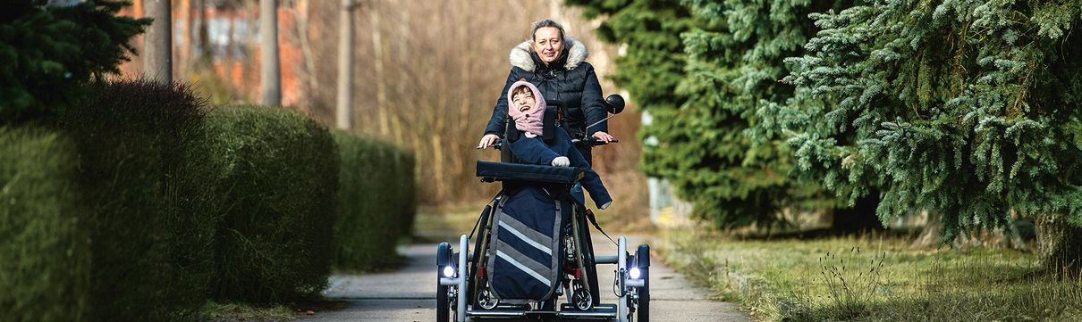Eine Frau fährt mit einem Lastenrad, auf dem ein Kind im Rollstuhl sitzt und voller Freude lacht, auf einem Weg auf den Betrachter zu.