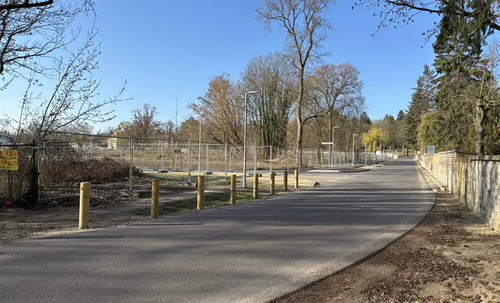 Ruhige asphaltierte Straße mit Pollern, rechts eine Mauer und links ein eingezäuntes Gelände mit Bäumen unter blauem Himmel.